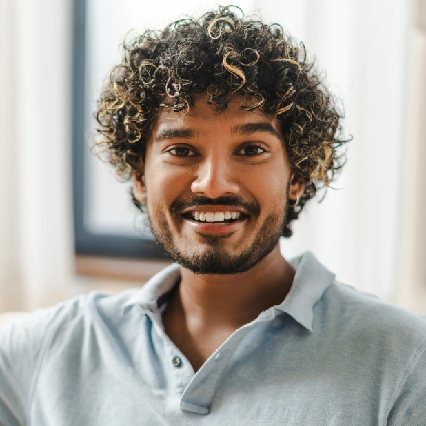 Smiling man with curly hair wearing light blue shirt in bright dental office setting.