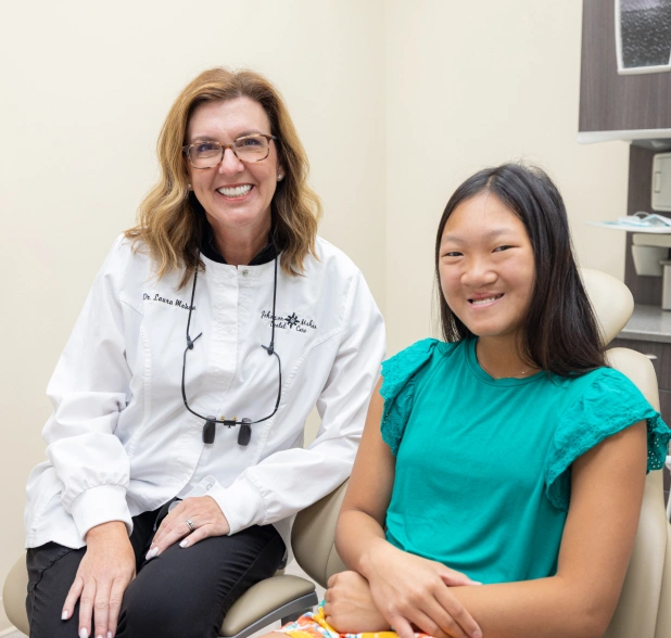 Two dental professionals smiling at camera in clinical office setting with shelving background.