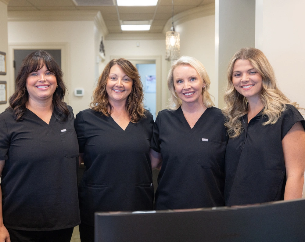 Four female dental staff members in black uniforms smiling in a modern dental office reception area.