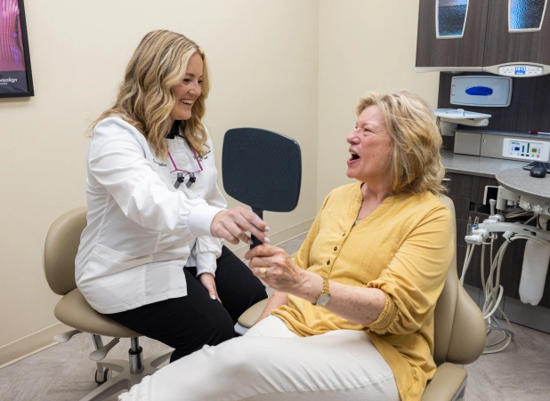 Two women shaking hands in a dental office examination room with medical equipment visible.