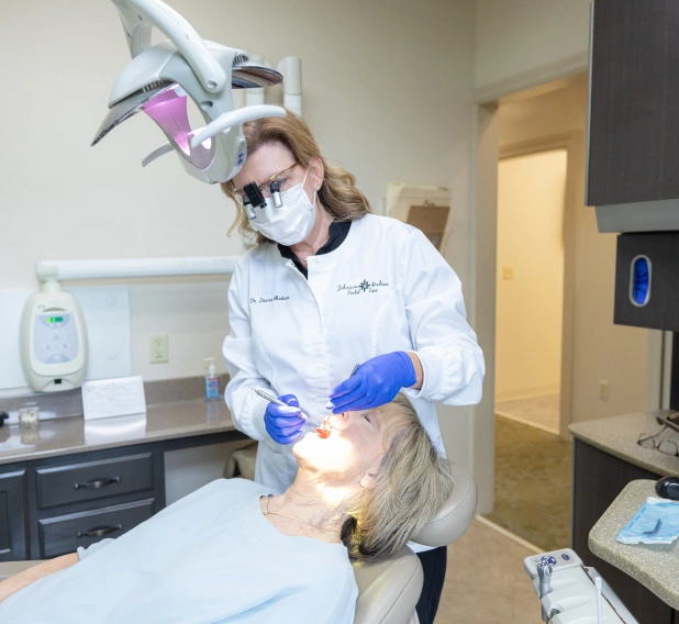 Dentist in white coat and blue gloves examining patient in dental office treatment room.