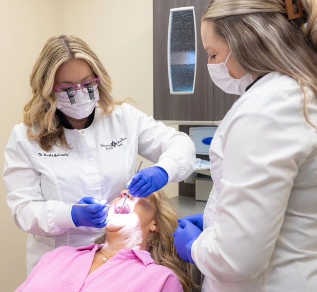 Two dental professionals in white coats and masks examine a young patient in a clinical setting.