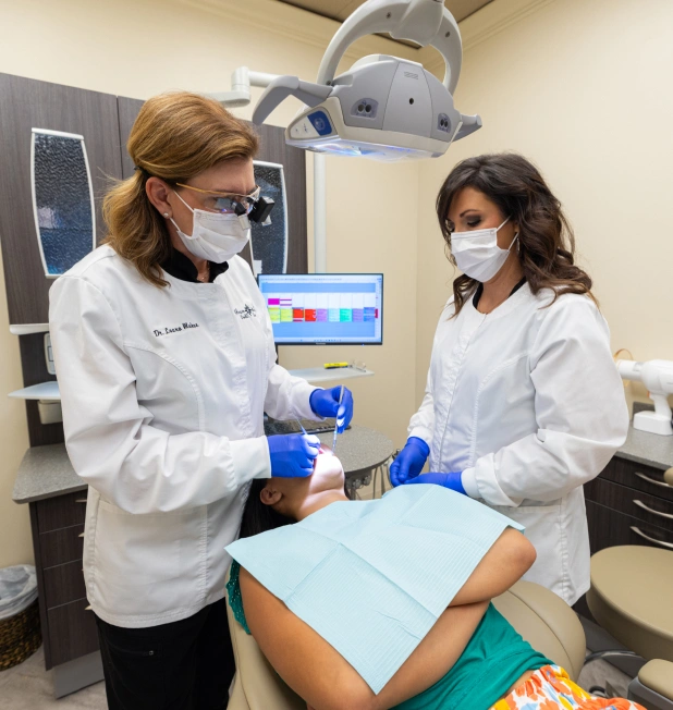Two dental professionals in white coats and masks prepare materials in a modern dental operatory.