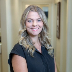 Professional headshot of smiling woman with blonde wavy hair in black shirt, dental office setting.