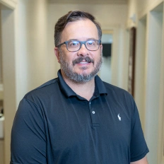 Man wearing glasses and dark polo shirt smiling in dental office hallway.