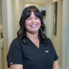 Professional dental staff member smiling in clinical office setting wearing black scrubs.