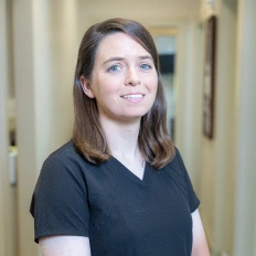 Woman in black scrubs smiling in dental office hallway setting.
