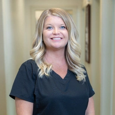 Blonde woman smiling at camera wearing black scrubs in dental office hallway.