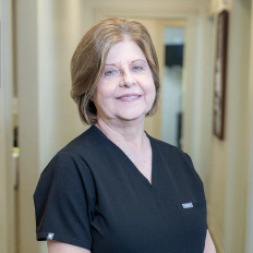 Female dental care professional wearing black scrubs smiling in clinical hallway setting.