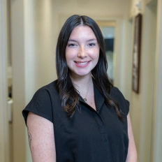 Professional woman smiling at camera in dental office hallway wearing black shirt.