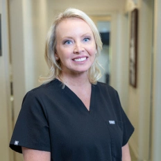 Professional female dental staff member in black scrubs smiling in clinical hallway.