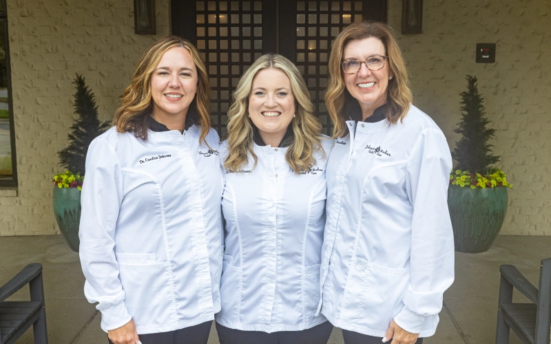 Three female dental professionals in white coats smiling at dental office entrance.